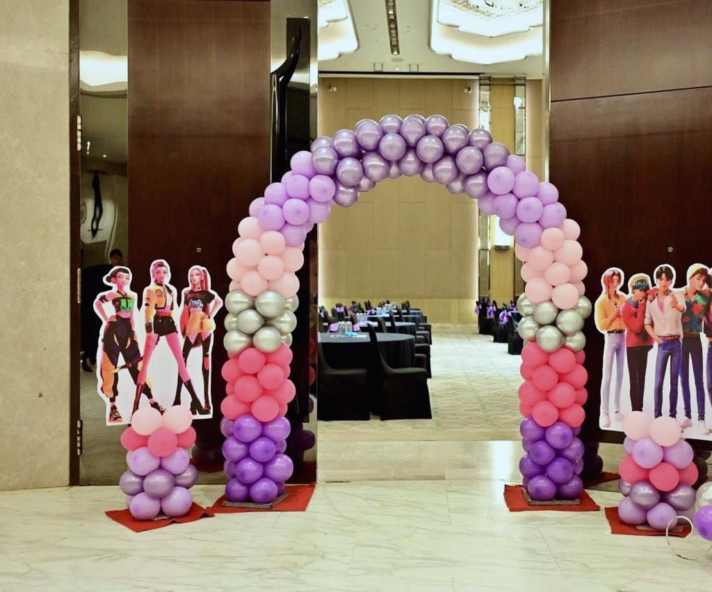 A pink, purple and silver balloon arch at a ballroom entrance, flanked by life-size K-pop cut-outs