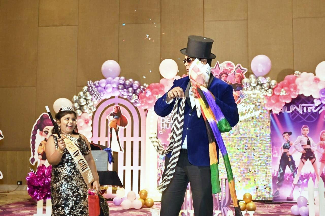 A magician in a top hat pulling rainbow silks from a newspaper as the birthday girl watches