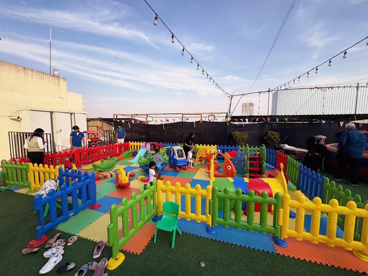 A rooftop soft play zone with slides, see-saws and colourful fencing under festoon lights